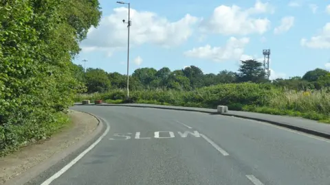 Robin Webster/Geograph This picture has been taken of a bend in Parsonage Road. "Slow" has been painted in white on the road surface. It is a sunny day. Bushes are on either side of the road.