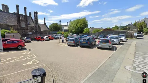 A car park is seen with a number of vehicles and buildings in the background on a sunny day