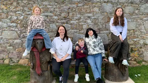 BBC A family photo with two young girls, two women and a young boy sitting on a wooden bench. 