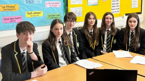 Ysgol Penrhyn Dewi Six Year 9 pupils are sitting around two desks put together. They all wear a white shirt, a navy, white and gold striped tie, navy blazer with gold edging on the collar and a school badge on their left. They are all white. The four girls all have long brown hair and the boys have short brown hair. Behind them on pinboards are various phrases in Welsh cut out into quote marks on brightly coloured laminated card.