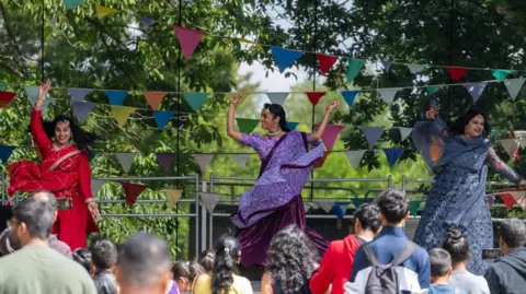 Ipswich Borough Council Two women in purple saree dresses dance on stage with their arms in the air. A crowd watches them. Bunting has been placed across the back of the stage.