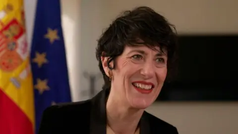 Juan Dominguez Elma Saiz, Minister for Inclusion, Social Security and Immigration, smiles in front of Spanish and EU flags