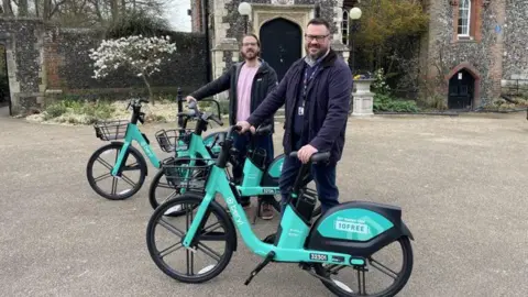 Two men stood next to three turquoise e-bikes.