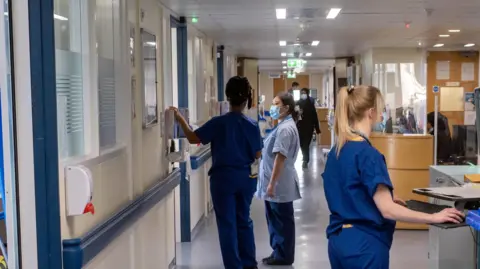 Nurses in scrubs each do different tasks in a hospital corridor, one looks at a computer, two others speak. 