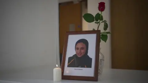 A framed headshot photo of Sara El-Khawas wearing a hijab. In front is a burning white candle, behind is a glass vase containing a single red rose.