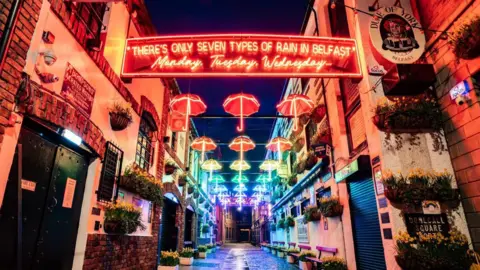 PA Media Different coloured umbrellas hanging on lines above a street. There are multiple signs and images on the walls of buildings surrounding the umbrellas. There is also a neon sign that reads '"There's only seven types of rain in Belfast" Monday, Tuesday, Wednesday...' in red. 