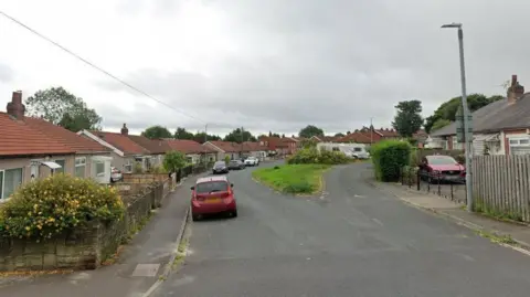 A road with single storey homes on both sides. The road forks and there is a grass verge in the middle. Two caravans are parked on the verge. Cars are parked on the edge of the street.