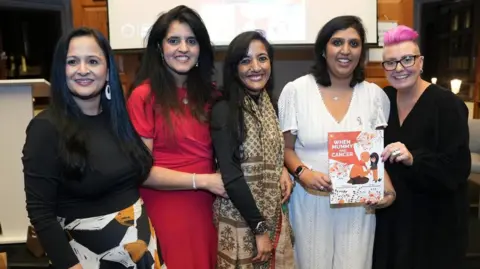 Author in Me Five women standing in front of a screen, smiling and looking towards the camera. The woman on the far left is looking slightly away, with long dark hair, a skirt and black top, the next woman long is wearing a red dress, and has long dark hair, the woman in the middle has on a long silk scarf and dark top, with long dark hair, Priyanka, is wearing a white lace all in one trouser outfit, holding her book, and the woman on the right has short pink hair, is also holding the book and is wearing all black. 