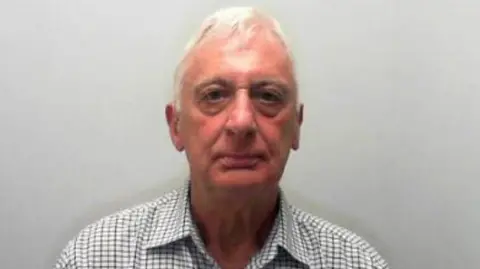 North Yorkshire Police Man with white hair wearing a checked shirt looks into a camera.