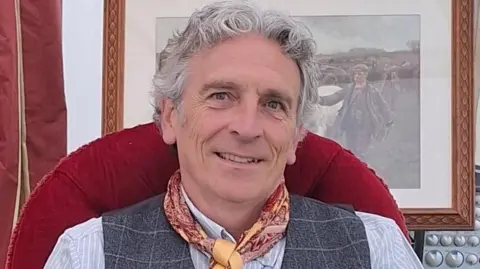 Handout A middle-aged man sits smiling on a red velvet chair. He wears a shirt, waistcoat and scarf tied around his neck. 