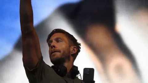 PA Media Calvin Harris wearing headphones around his neck raises his arm in the air on stage 