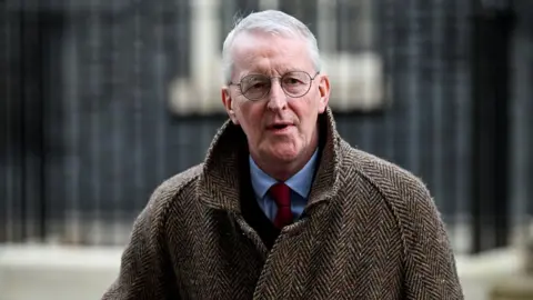 Reuters The Secretary of State for Northern Ireland, Hilary Benn MP in a black suit and red tie, with a tweed coat over it. He is mid speech. He has white hair and glasses