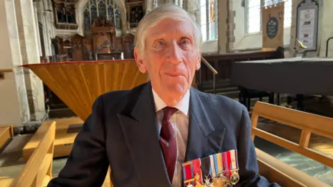 Major General Patrick Cordingley wearing a dark navy suit and a strip of colourful wartime medals on his lapel. He has white hair and blue eyes, and is standing near the front of a church, with a stained glass window and wooden pews in the background. 