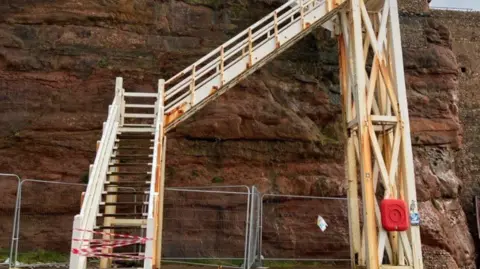 A view from the bottom of Jacob's Ladder steps in Sidmouth. The stairs are white and has a lot of rusting on it. There is a red/brown cliff behind it.