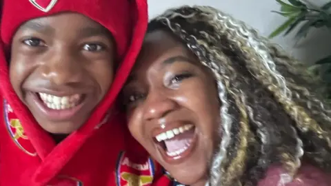 A young boy wears a red Arsenal hoodie and hat, next to a woman with multi-coloured hair. Both are smiling. 