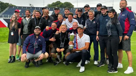 The Belfry British Masters golfers gather on The Belfry's course with Anna Nilsson. There are more than 20 people in the photo with one man holding a trophy.