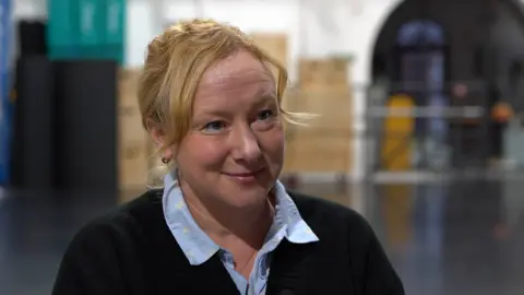 Sarah Worth, the co-founder of Highly Sprung Performance in Coventry. She is wearing a blue shirt under a black V-necked jumper and has blonde hair tied back