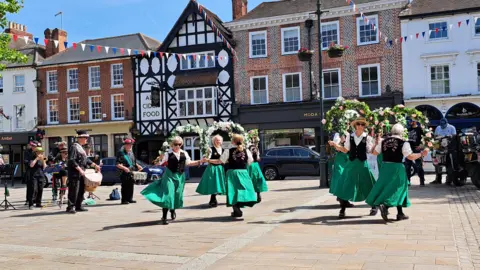 Fiddlesticks A troupe of eight female clog dancers on a paved area in a town centre, with shops in the background. They are wearing calf-length green skirts, black waistcoats and white, short-sleeved collared shirts. They are jigging and holding garlands. They have two male drummers and a couple of other musicians with them. It is a sunny day.