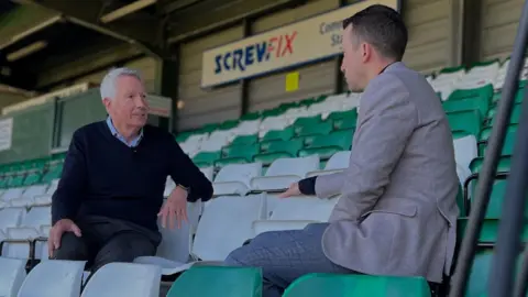 Yeovil Town FC Two men are sitting facing each other on the stands at a football arena. The seats are grey and green.