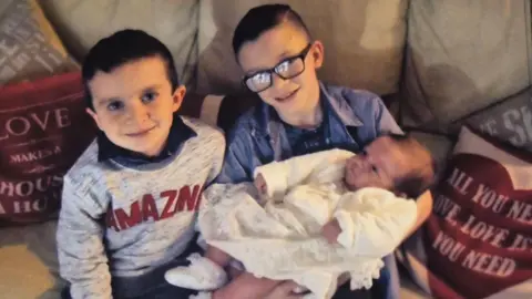 Louise James Evan and Mark with their sister Rioghnach  . Evan is wearing a grey sweater with large red letters spelling out AMAZING. He has short black hair. Next to him Mark has black square-rimmed glasses and is wearing a blue shirt. Both boys are sitting on a tan sofa with pink cushions. Mark is holding Rioghnach who is wearing a white dress and cardigan.