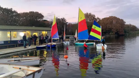 Sailing boats on the lake, sails flag colours are blue, yellow and red with the boathouse and parents in the background.