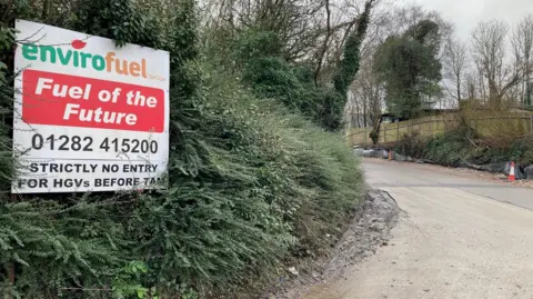 A sign with green, orange and black lettering reads: "Envirofuel - Fuel for the future" surrounded by green bushes next to the entrance to the site.