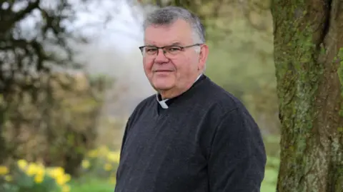 Mark Christian is pictured wearing a dark grey V-neck jumper with a white dog collar. He is standing outside, next to a tree, on a cloudy day. He has short grey hair and is wearing black-rimmed glasses. He is looking at the camera in a slightly pensive way.