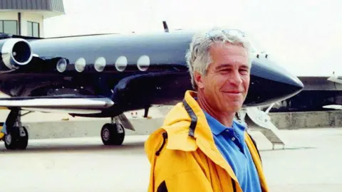Jeffrey Epstein, a man with grey hair wearing a bright blue polo shirt and an orange anorak, smiling broadly as he stands in front of his private plane - a black jet with chrome detailing on the wings and around the engines, with five porthole-style windows visible on the right-hand side.