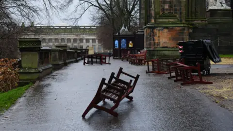 PA Media Benches in Princes Street Gardens in Edinburgh. They are next to the Scott Monument. All are brown and have been blown over. One is in the middle of the picture in the foreground and is upside down. Others have been blown over to the side of the image.