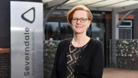 Severndale Academy A woman with short red hair and glasses. She is smiling and wearing a black cardigan on top of a black patterned shirt.