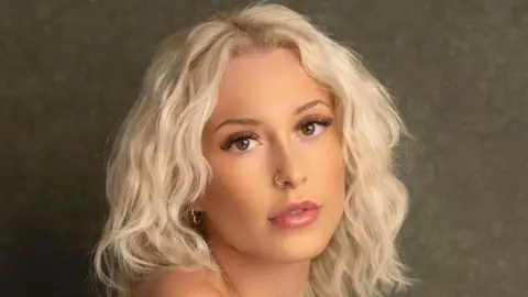 A close-up headshot of a woman with platinum blonde curly hair. She looks at the camera in front of a dark green background.