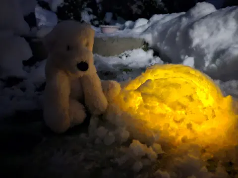 Nikki Swinburn A stuffed toy bear sitting outside in the snow, next to a small igloo lit-up with lights.