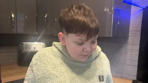 Family handout Rory, a boy with sort brown hair stands in the kitchen of his home. He is wearing a grey fleece top. He is looking at the floor. 