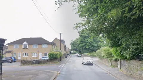 A road showing a junction with another road. There is a car on the road and houses and greenery in the background. 
