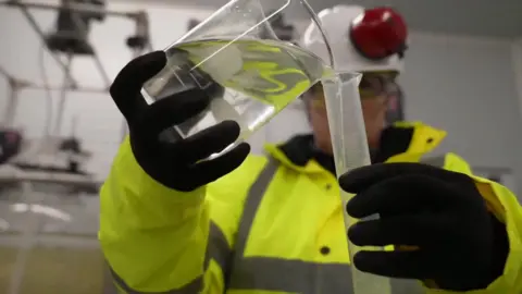 A picture of someone pouring a liquid from a glass into a tube. They are wearing protective glasses and headgear - alongside a green jacket and black gloves.