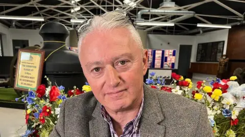 Qays Najm/BBC David Hadjicostas smiles at the camera, seated in front of some multi-coloured flowers. He is wearing a tweed jacket and plaid shirt.