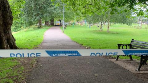 French Weir Park in Taunton. Police tape can be seen across the frame, and trees, grass and a play park in the distance. 