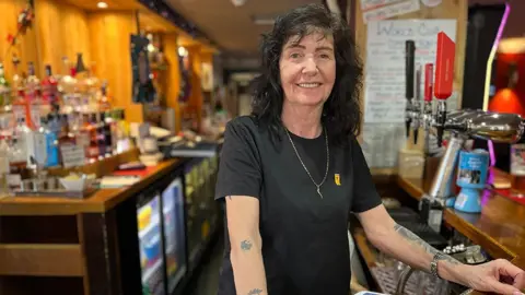 Carol Graham, who has black curly hair, smiles while standing behind a bar. She is wearing a black t-shirt and has tattoos on her forearms.