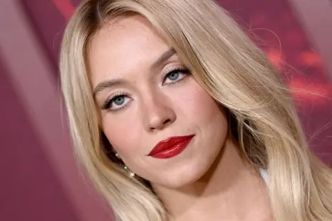 A close-up shot of Sydney Sweeney on a red carpet. She looks glamourous, with her blonde hair in a sleek, centre-parted style and a bold red lip.