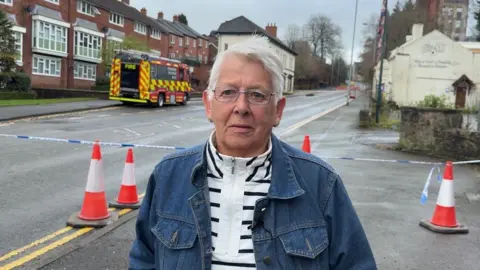 Dianne Donohue, a woman wearing a black and white top, and a denim jacket, is standing by a police cordon. There is a fire engine and a set of orange traffic cones behind her. There are buildings either side of the road behind.