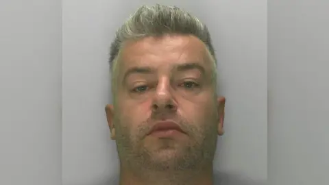 Mugshot of a man in his early 40s with grey hair which is short on the sides and forms a small quiff on top