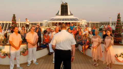 Villa Vie Residences A captain facing away from the camera officiates a wedding on the deck of a cruise ship. The couple are in front of him. Bridesmaids stand to the right and grooomsmen to the left. Guests stand behind the couple.
