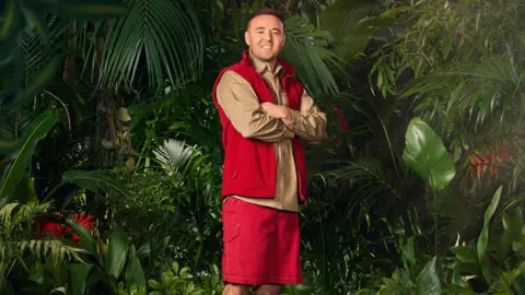 ITV Alan Halsall wearing his jungle outfit of red shorts, an earth-toned shirt and red gilet, in a jungle scene surrounded by plants