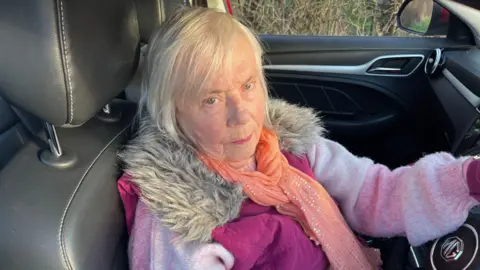 John Devine/BBC Sally Belcher-Greenhalgh has grey hair swept across her head, she is wearing a woolly pink jumper with a darker pink body warmer, with an orange chiffon scarf around her neck, she is sitting in the drivers seat of a car and looks fed up.