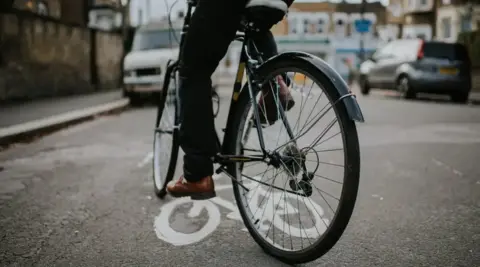 Getty Images An image of a cyclist on a bike. The image is taken from behind them and only the back of their lower legs straddling the bicycle in the road are visible. 