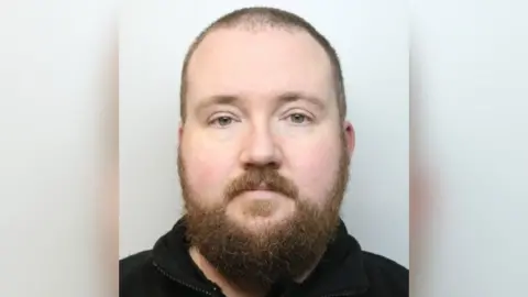 A police photo shows a man with a large beard and wearing a dark fleece