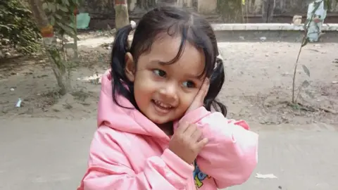  Al Amin A young girl wearing a pink hooded jumper with her hair in pig-tails holds a hand against her face, smiling