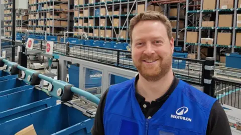 Alastair Bell Alastair Bell smiling and looking down the camera. He is wearing a blue zipped gilet with 'Decathlon' and a logo on one shoulder. Underneath is a black t-shirt. He is standing inside a warehouse, with blue plastic and cardboard boxes arranged behind him. There are a large number of shelves holding the boxes arranged behind him. 
