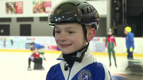 A nine-year-old boy smiles while standing in front of an ice rink. He is wearing a black helmet and blue and white body suit with a yellow rose embroidered on the right side.