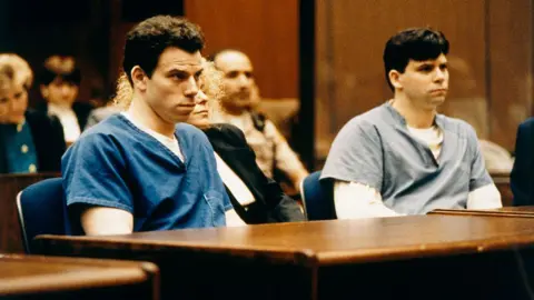 The Menendez brothers sit in prison suits inside a courtroom. In between them, their defence attorney sits at the wooden table. 